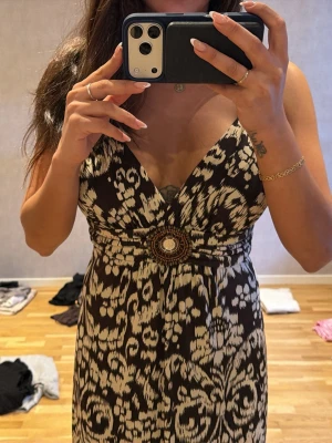 Mönstrad långklänning med v-ringning - Snygg långklänning med djup v-ringning och bred midjedetalj med dekorativt spänne. Klänningen har ett svart och beige mönster och är ärmlös med smala axelband. Perfekt för sommarens alla tillfällen. Köpt i Cannes.
