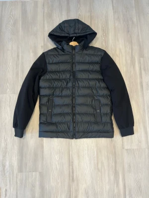 Svart hybridjacka - Säljer en svart pufferjacka med avtagbar huva och hel dragkedja framtill. Jackan har quiltad kropp, släta ärmar och två fickor med knappstängning. Perfekt för kalla dagar och riktigt snygg streetstil.