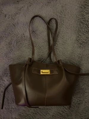 Mörkbrun handväska med gulddetalj - Snygg mörkbrun handväska i slätt konstläder med långa axelband och guldfärgad metallspänne framtill. 