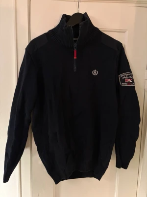 Mörkblå stickad tröja Henri Lloyd - Snygg mörkblå stickad tröja från Henri Lloyd med halv dragkedja och hög krage. Tröjan har patch med logga på ena ärmen och liten broderad logga på bröstet. Ribbstickade muddar och axelpartier i förstärkt material. Perfekt för kyliga dagar.