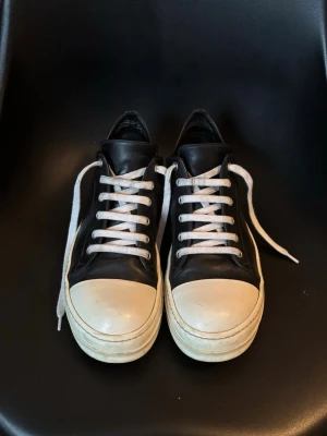 Rick Owens mainline ramones - Rick Owens mainline ramones i svart läder. Strl 42. Ingen låda. Använda men fortfarande i väldigt bra skick. Insidan av höger sula har kommit i kontakt med ett varmt avgasrör som lämnat ett litet märke men det är knappt synligt (se sista bilden). Efter åratal av slitage började lädret runt fotöppningen spricka (1 mm) och har reparerats nästan osynligt av en skomakare för att förhindra ytterligare sprickor. Så allt som allt i gott skick. 