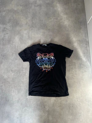 Svart Kenzo t-shirt med tigertryck - Snygg svart t-shirt från Kenzo med ikoniskt tigertryck i flera färger på bröstet. Klassisk passform och rund halsringning. Perfekt för dig som vill sticka ut med en färgstark detalj. Tillverkad i mjuk bomull för skön känsla.