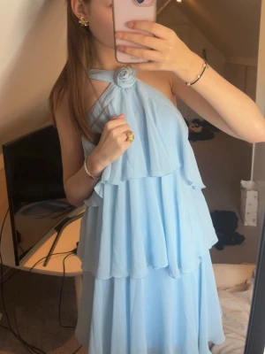 Ljusblå volangklänning med rosdetalj - Supersöt ljusblå klänning med volanger och halterneck med en rosdetalj vid halsen. Klänningen har flera lager och är ärmlös, perfekt för sommarens alla tillfällen. Materialet känns lätt och luftigt, troligtvis polyester eller liknande. Prislapp kvar!! Fint skick och aldrig använd!🦋