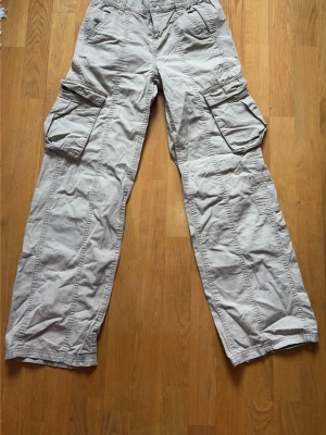 Ljusgrå cargobyxor med stora fickor - Säljer ett par ljusgrå cargobyxor med bred passform och flera stora fickor på sidorna. Byxorna har elastisk midja och klassiska cargodetaljer. Perfekta för en avslappnad och trendig streetwear-look.