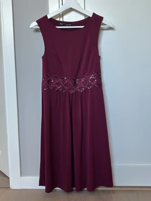 Vinröd klänning med spetsdetalj - Vinröd långklänning med breda axelband och vacker spetsdetalj med pärlor i midjan. Klänningen har en elegant, något utsvängd passform och är perfekt för speciella tillfällen. Materialet känns mjukt och följsamt.