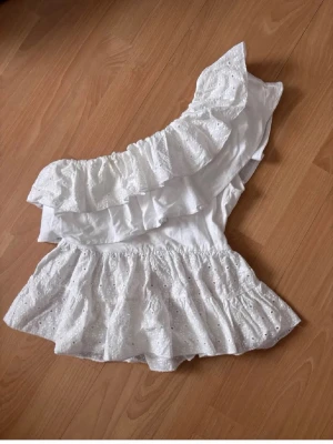 Zara blus  - Säljer en vit oneshoulder topp med stora volanger och broderade detaljer. Perfekt för dig som gillar en unik och feminin stil. Toppen är ärmlös och har en luftig passform med vackra spetsmönster i tyget. TROR S