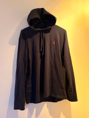 Marinblå hoodie från Ralph Lauren - Marinblå hoodie med huva och dragsko från Ralph Lauren. Klassisk design med lång ärm och den ikoniska röda logotypen broderad på bröstet. Tillverkad som en långärmad t shirt med huva. Perfekt för en avslappnad och stilren look. 