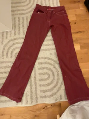 Vinröda bootcut jeans - Säljer ett par vinröda bootcut jeans med klassisk femficksdesign och vita kontrastsömmar. Jeansen har normal midja och är tillverkade i ett mjukt denimtyg som ger en skön passform. Perfekta för dig som vill sticka ut med färg i vardagen.