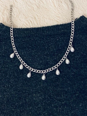 Silverfärgat halsband med pärldroppar - Snyggt halsband i silverfärgad metall med sju droppformade vita pärlor som hänger längs kedjan. Kedjan är grov och ger ett trendigt intryck, perfekt för att lyfta vilken outfit som helst. Märke saknas på halsbandet.