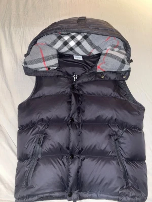 Burbbery jacka - Säljer en svart dunväst från Burberry med avtagbar huva och klassiskt rutigt foder i grått, vitt och rött. Västen har dragkedja och knappar framtill samt två fickor med dragkedja. Perfekt för lager på lager och riktigt snygg till höst och vinter. S/ mindre snabbaffär funkar också 