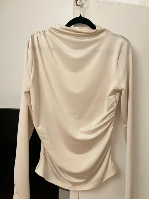 Beige långärmad topp från H&M - Säljer en beige långärmad topp från H&M med draperad effekt på sidan och hög hals. Toppen har en mjuk, stretchig känsla och sitter snyggt figurnära. Perfekt att styla med jeans eller kjol för en clean look.