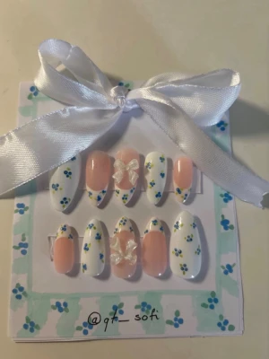 Press-on naglar med blå blommor och rosetter - Söta press-on naglar i mandelform med vit och ljusrosa bas, dekorerade med små blå och gula blommor samt två genomskinliga 3D-rosetter. Perfekt för dig som vill ha en gullig och unik nagelstil. Kommer i ett set om tio naglar.