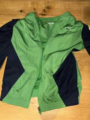 Grön och marinblå vindjacka Lacoste. Köpte för 650kr, oanvänd. - Sportig vindjacka från Lacoste i grönt och marinblått med dragkedja framtill och elastiska muddar. Jackan har klassisk Lacoste-logga på bröstet och är tillverkad i lätt, vindtätt material. Perfekt för en aktiv stil.