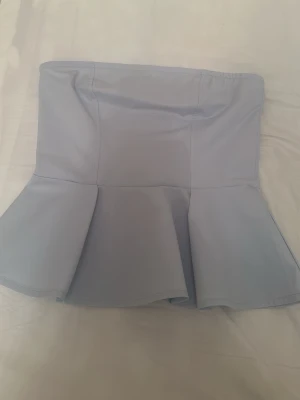 Ljusblå peplum tubtopp - Snygg ljusblå tubtopp med peplumvolang nertill. Toppen är axelbandslös och har en figurnära passform upptill som släpper ut i en volang. Perfekt för dig som vill ha en trendig och stilren look.