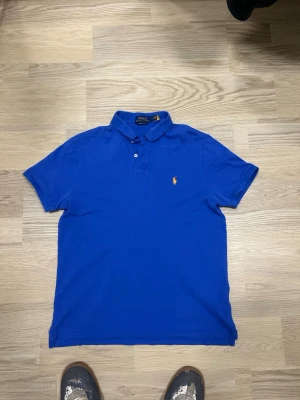 Blå pikétröja från Polo Ralph Lauren - Snygg blå pikétröja från Polo Ralph Lauren med klassisk krage, korta ärmar och knappar framtill. Tröjan har den ikoniska broderade loggan på bröstet och är i bomull med slim fit-passning.