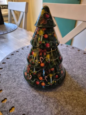 Julgranformad plåtburk - Säljer en julgranformad burk i plåt med färgglada julmotiv som polkagrisar, apelsiner och kottar. Burken är grön med detaljer i rött, gult och vitt och kan delas i flera delar för förvaring av småsaker eller godis.