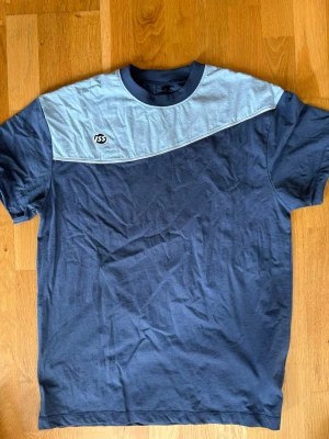 Blå och ljusblå t-shirt från ISS - Snygg t-shirt från ISS i två nyanser av blått. T-shirten har en mörkblå bas med ljusblå panel upptill och en liten ISS-logga på bröstet. Klassisk rund halsringning och korta ärmar. Perfekt för en avslappnad stil.