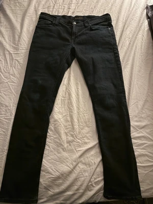 Svarta jeans från Nudie Jeans Co tight long john - Säljer ett par svarta jeans från Nudie Jeans Co med klassisk femficksdesign och smal passform. Jeansen har knappgylf och subtila detaljer, perfekta för en stilren look. Materialet är mjukt och stretchigt för extra komfort.