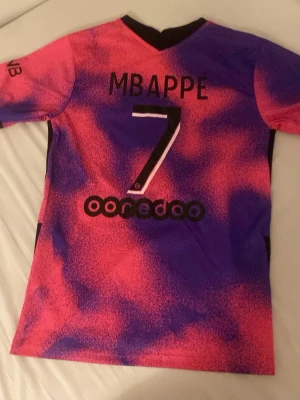 PSG Mbappé matchtröja rosa/lila - Säljer en PSG matchtröja i samarbete med Jordan, med Mbappé och nummer 7 på ryggen. Tröjan är rosa och lila med svarta detaljer, kort ärm och rund hals. Materialet är lätt och andas, perfekt för fotboll eller träning. 100% äkta. Köpt i Paris 