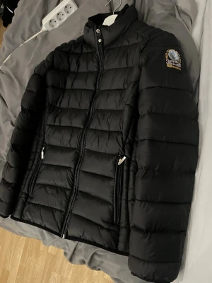 Svart pufferjacka från Parajumpers - Svart pufferjacka från Parajumpers med hög krage, dragkedja framtill och två fickor med dragkedja. Jackan har ett klassiskt quiltat mönster och en patch med Parajumpers-logga på ena ärmen. Perfekt för kalla dagar och riktigt snygg streetstil.