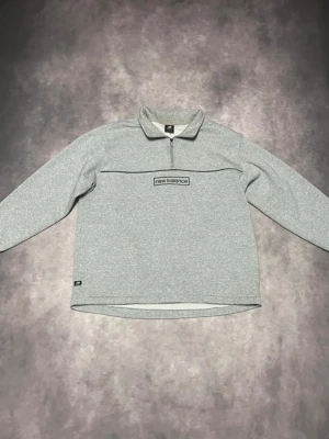 Grå halvzip tröja från New Balance - Snygg grå långärmad tröja från New Balance med halv dragkedja och broderad logga på bröstet. Tröjan har en avslappnad passform och är tillverkad i mjukt material, perfekt för en sportig och casual stil.