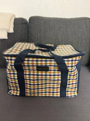 Rutig resväska från Discover - Färgglad rutig resväska från Discover med blå, gula och röda rutor. Väskan har två kraftiga blå handtag och dragkedja upptill. Tillverkad i slitstark textil och har en mjuk, boxig form som gör den enkel att packa.