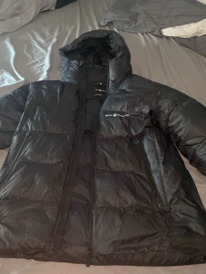 Svart pufferjacka Sail Racing - Säljer en svart pufferjacka från Sail Racing med huva och dragkedja framtill. Jackan har två snedställda fickor med dragkedja och logga på bröstet. Perfekt för kalla vinterdagar och riktigt skön att bära. Klassisk boxig passform och fluffigt material.