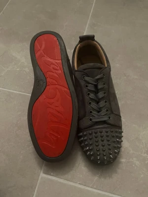 Svarta Louboutin sneakers med nitar - Säljer ett par svarta sneakers från Christian Louboutin som knappt är använda och de är ny tvättade. med klassisk röd sula och coola nitar på tån. Skorna har snörning och är gjorda i mocka för en lyxig känsla. Perfekta för dig som vill sticka ut med din stil. DOM ÄR I STORLEK 39-41