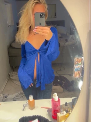 Blå omlottopp med knyt framtill - Snygg blå omlottopp med knyt framtill och djup v-ringning. Toppen har långa ärmar och är croppad, vilket ger en trendig och avslappnad look. Perfekt för dig som vill sticka ut med en färgstark och unik topp.