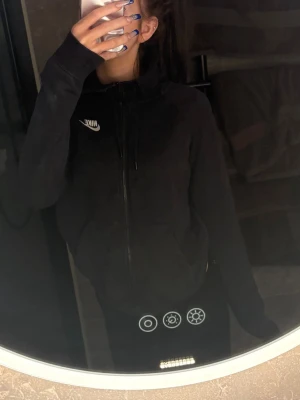 Svart Nike zip hoodie med huva - Säljer en svart zip hoodie från Nike med vit logga på bröstet. Hoodien har dragkedja hela vägen, två fickor framtill och justerbar huva med snören. Perfekt för chill dagar eller när du vill ha en clean och enkel look.