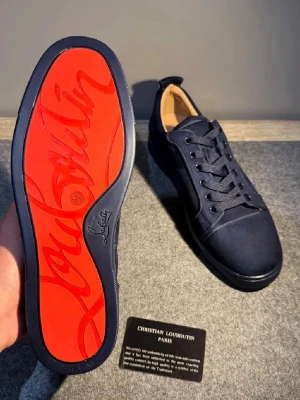 Christian Louboutin svarta sneakers - Snygga svarta sneakers från Christian Louboutin med ikonisk röd sula och klassisk låg profil. Skorna har en stilren design med svarta snören och ovandel i textil. Perfekt för dig som vill ha en exklusiv och trendig look.