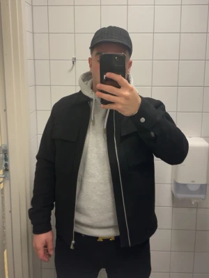 Svart overshirt med fickor från Jack and Joans  - Säljer en svart overshirt med klassisk krage och två bröstfickor med lock. Jackan har raka ärmar med knapp vid ärmslut och dragkedja framtill. Perfekt att bära över en hoodie för en avslappnad look.