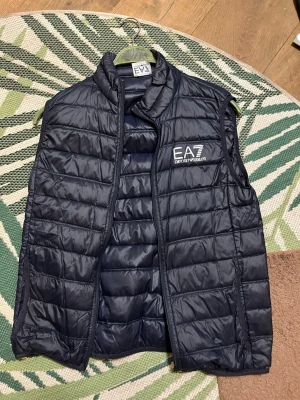 Svart dunväst från EA7 Emporio Armani - Storlek S Snygg svart dunväst från EA7 Emporio Armani med quiltad design och hög krage. Västen har dragkedja framtill och logga tryckt på bröstet. Perfekt för lager-på-lager och streetstyle.