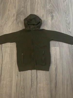 Olivgrön vindjacka med huva - Snygg olivgrön vindjacka med huva och dragkedja framtill. Jackan har två fickor med dragkedja och ribbad struktur på framsidan. Perfekt för blåsiga dagar och enkel att matcha med olika outfits. Indicode, bra skick, Large fits Medium.