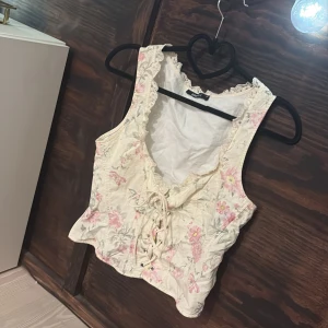 Blommig korsettopp från Gina Tricot - Säljer trendig korsettopp från Gina Tricot i cremevitt tyg med broderade rosa blommor. Toppen har snörning framtill, spetsdetaljer vid halsringningen och är ärmlös. Perfekt för dig som vill ha en söt och trendig look. Använd en gång! 