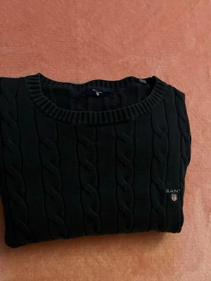 Svart kabelstickad tröja från Gant - Kabelstickad svart tröja från Gant med rund halsringning och broderad logotyp på bröstet. Klassisk design med ribbade muddar och mjukt material som passar perfekt till jeans eller kjol.