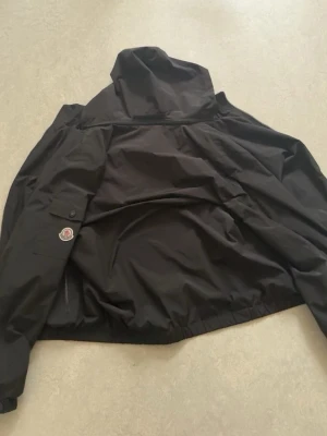 Svart vindjacka från Moncler - Snygg svart vindjacka från Moncler med huva och dragkedja framtill. Jackan har två fickor med dragkedja och Moncler-logga på ärmen. Tillverkad i lätt och slitstarkt material, perfekt för blåsiga dagar.