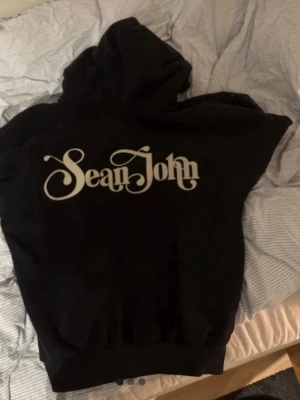 Svart hoodie från Sean John - Svart hoodie från Sean John med stor vit logga på ryggen och mindre logga framtill. Klassisk modell med dragkedja, huva och fickor. Perfekt för en avslappnad streetwear-look. Tillverkad i mjukt material som känns skönt mot huden.