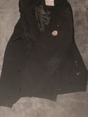 Svart vindjacka från Moncler - Snygg svart vindjacka från Moncler med klassisk logotyp på bröstet. Jackan har dragkedja, elastiska muddar och fickor med dragkedja. Tillverkad i lätt och slitstarkt material som passar perfekt för blåsiga dagar.