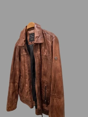 Brun skinnjacka från Saki - Snygg brun skinnjacka från Saki med klassisk krage och dragkedja framtill. Jackan har flera fickor med dragkedja och en cool vintage-look. Perfekt för dig som vill ha en tidlös och stilren jacka i äkta skinn.