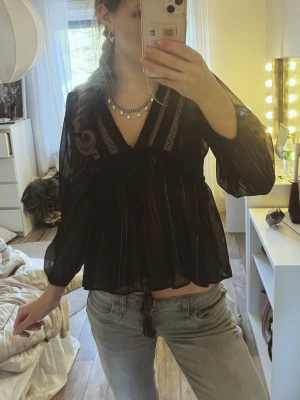 Svart blus - Snygg svart blus med transparent material och subtila ränder. Blusen har v-ringning, spetsdetaljer och knytband runt om. Guld detaljer på