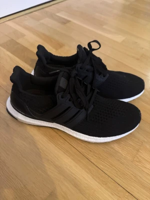 Adidas Ultra Boost svarta sneakers - Svarta Adidas Ultra Boost sneakers med stickad ovandel och klassiska tre ränder på sidan. Skorna har en vit Boost-sula för extra komfort och svart hälkappa. Perfekta för dig som gillar sportig och stilren look.