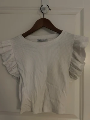 Vit ribbad topp med volangärmar från Zara - Säljer en vit ribbad topp från Zara i storlek M, kan även används som S. Toppen har breda, ärmlösa axlar med snygga volangdetaljer som ger en cool och trendig look. Materialet är mjukt och stretchigt, perfekt för att lyfta din outfit med lite extra stil. Säljer då den ej kommer till användning