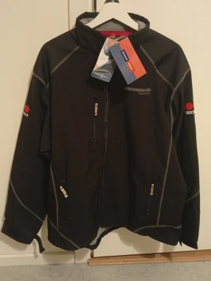 Svart softshelljacka från Fristads - Svart softshelljacka från Fristads med Securitas-tryck på ärmen och bröstet. Jackan har dragkedjor framtill och på fickorna, samt grå kontrastsömmar. Perfekt för dig som vill ha en funktionell och stilren jacka med sportig känsla.