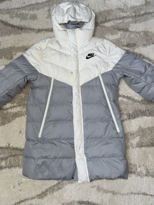 Lång nike jacka  - Snygg dunjacka från Nike i grått och vitt med huva och dragkedja framtill. Jackan har två stora fickor med dragkedja och svart Nike-logga på bröstet. Perfekt för kalla dagar och riktigt skön att bära.