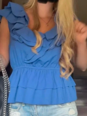 Blå volangblus från H&M - Supersöt blå blus från H&M med flera volangdetaljer och rund halsringning. Blusen har ärmlös design och smickrande passform med resår i midjan. 