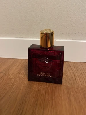 Versace Eros Flame EDP 2ml sample - Versace Eros Flame Eau de Parfum i en elegant mörkröd glasflaska med guldigt lock. Flaskan har en fyrkantig form och är dekorerad med Versaces ikoniska Medusa-huvud på framsidan. Perfekt för dig som gillar lyxiga och intensiva dofter.