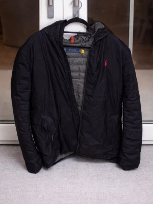 Mörkblå packbar dunjacka Polo Ralph Lauren - Snygg mörkblå dunjacka från Polo Ralph Lauren med röd logga på bröstet och diskret 'PACKABLE'-text. Jackan är lätt, quiltad och har en grå insida. Perfekt för kalla vinterdagar och enkel att packa ner i väskan.