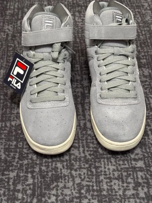 Gråa Fila high top sneakers - Stilrena gråa sneakers från Fila med högt skaft och kardborreband upptill. Skorna har mockaliknande ovandel, klassiska snörningar och vit sula. Perfekta för dig som gillar en clean och sportig look. Fila-logga på plösen och sidan.