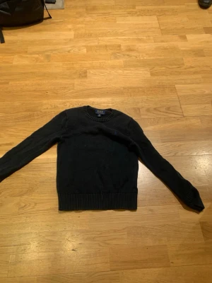 Svart stickad tröja från Polo Ralph Lauren - Klassisk svart stickad tröja från Polo Ralph Lauren med rund halsringning och blå broderad logga på bröstet. Tröjan har långa ärmar och ribbstickade muddar vid ärmslut och nederkant. Perfekt för en stilren look.